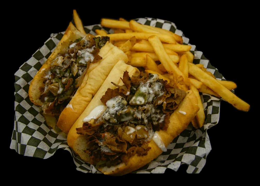 A Philly cheesesteak with ribeye, peppers and onions next to fries in a checkered basket